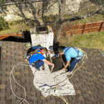 Two technicians performing chimney maintenance on a rooftop, using safety harnesses and equipment, with a chimney and tree in the background, related to Doodlebuggers Fireplace services.