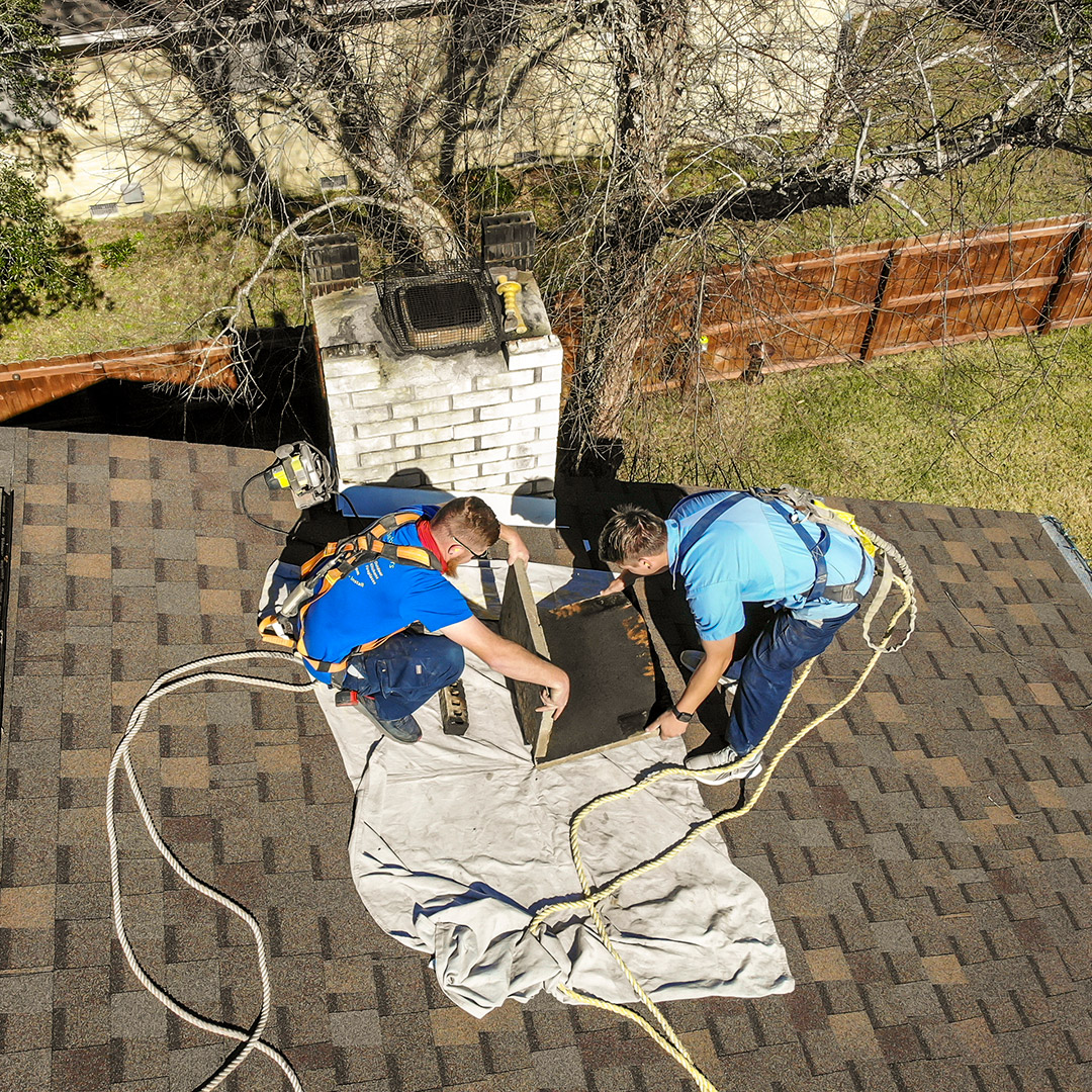 Two technicians working on a roof near a chimney, preparing for chimney repair, with safety harnesses and tools visible, emphasizing professional chimney maintenance services.